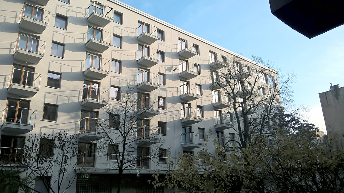 Fasada nowoczesnego budynku mieszkalnego z licznymi balkonami o szklanych balustradach, widziana z dołu pod kątem, z drzewami na pierwszym planie.