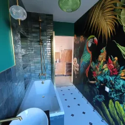 Nowoczesna łazienka z wanną, armaturą w kolorze złota i ścianą z motywem tropikalnym z papugą, oświetlona wiszącą lampą.