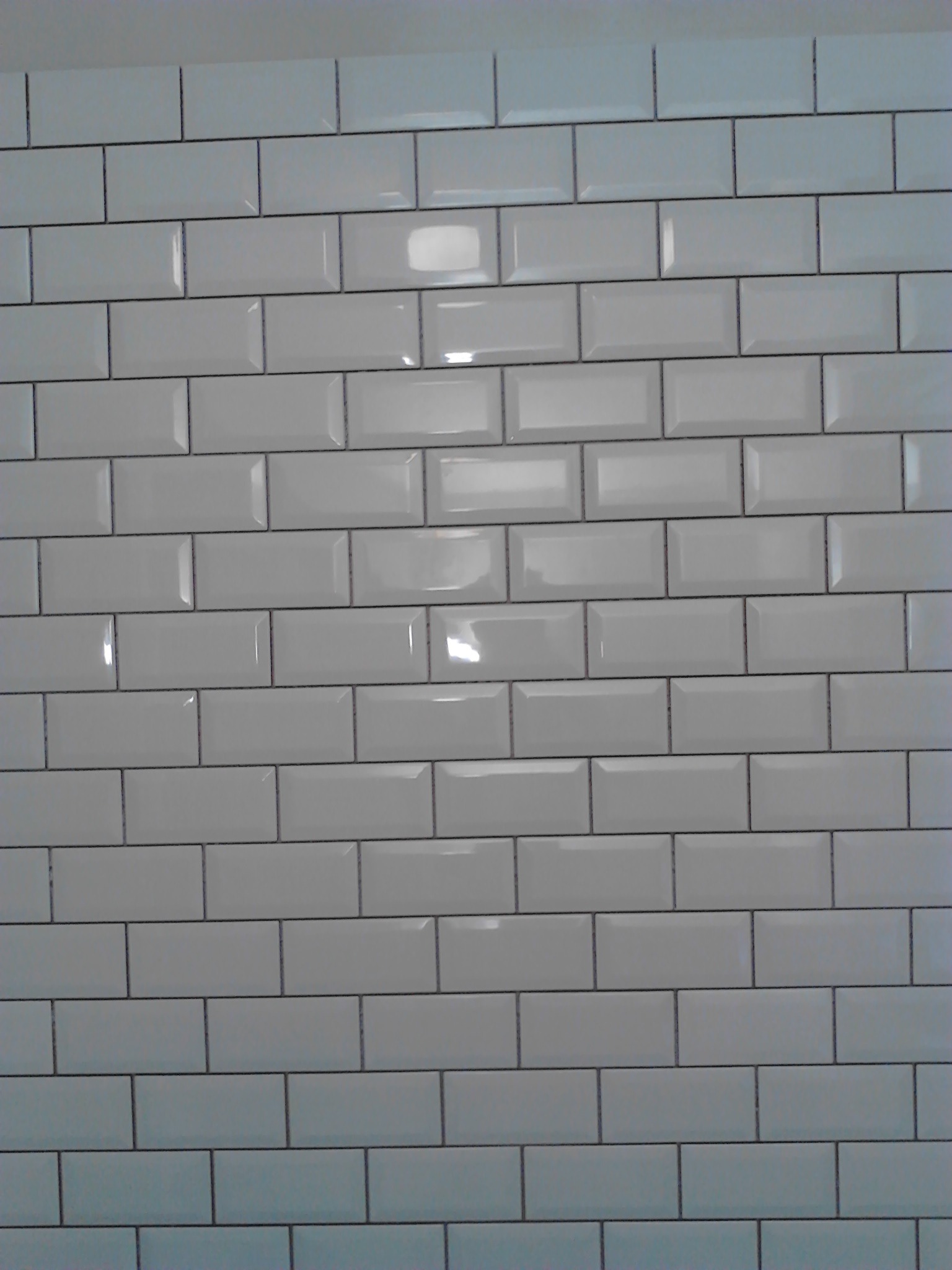 Ściana wyłożona białymi, błyszczącymi płytkami typu metro z ciemną fugą, wykonana w Toruniu. Widoczne odbicie światła na powierzchni płytek.