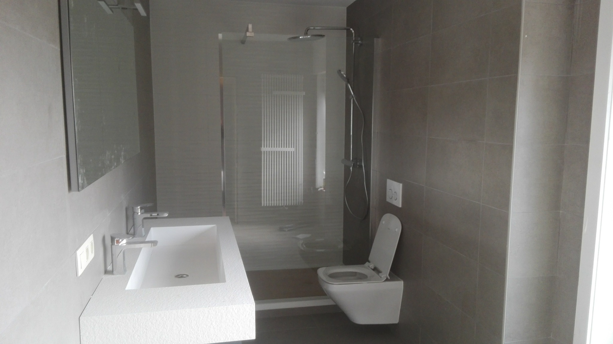 Nowoczesna łazienka z szarymi płytkami, umywalką z białego kamienia, kabiną prysznicową ze szklaną ścianką i podwieszaną toaletą z otwartą klapą.