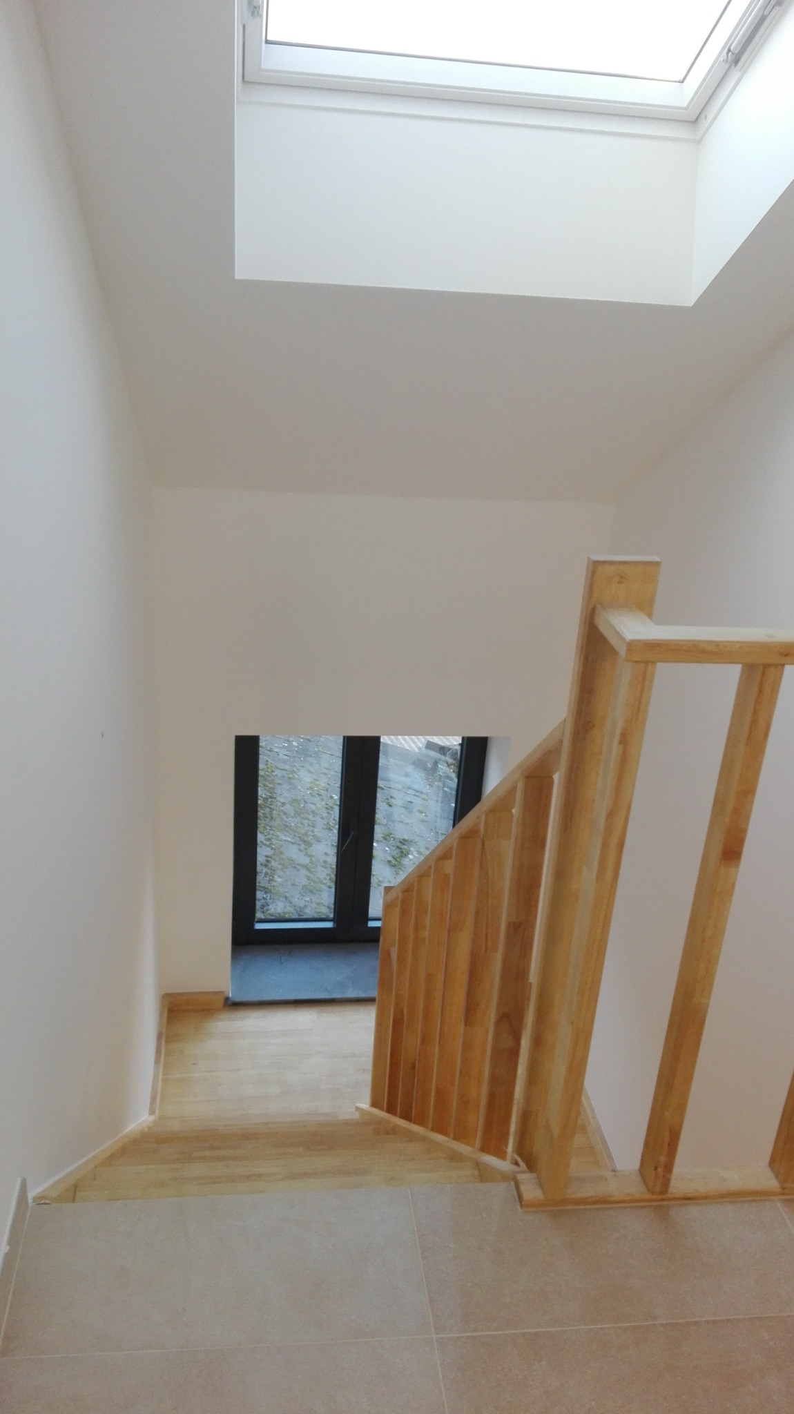 Jasne, świeżo pomalowane wnętrze klatki schodowej z drewnianą balustradą, płytkami podłogowymi i oknem dachowym, widok z góry na schody prowadzące do przeszklonych drzwi balkonowych.