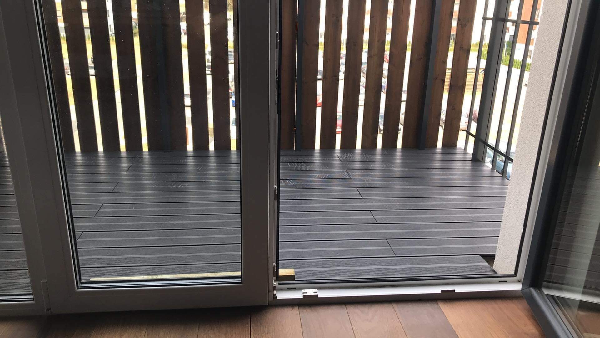 Nowoczesny balkon z szarą, kompozytową podłogą, drewnianą balustradą z pionowych desek i rozsuwanymi drzwiami balkonowymi.