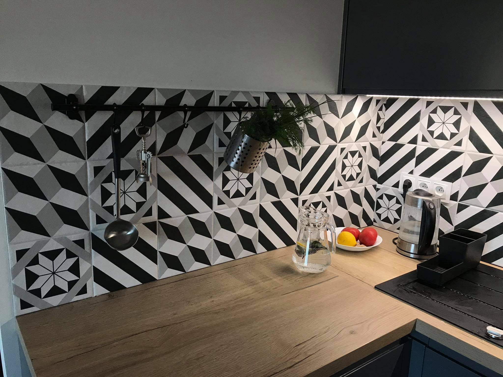 Kuchnia z czarno-białymi geometrycznymi płytkami na ścianie, drewniany blat, czarny reling z zawieszonymi akcesoriami kuchennymi i ziołami, szklany dzbanek z wodą i cytryną oraz czajnik elektryczny.