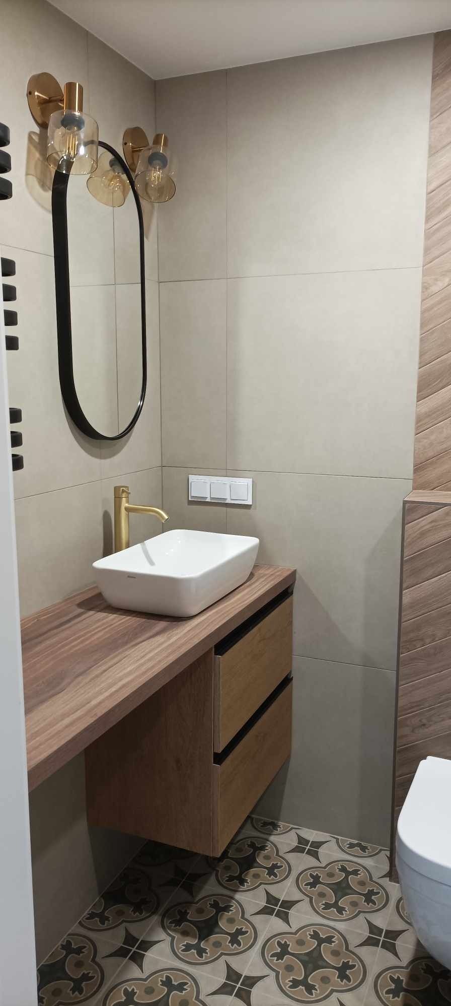 Nowoczesna łazienka z beżowymi płytkami, drewnianą szafką pod umywalką z szufladami, białym blatem i umywalką nablatową, złotą baterią, owalnym lustrem w czarnej ramie i wzorzystą podłogą.