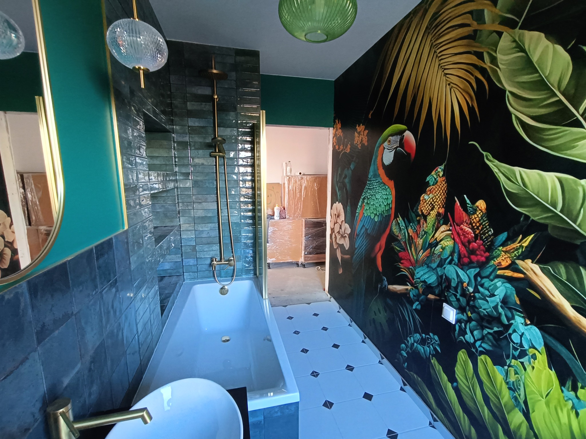 Nowoczesna łazienka z wanną, armaturą w kolorze złota i ścianą z motywem tropikalnym z papugą, oświetlona wiszącą lampą.