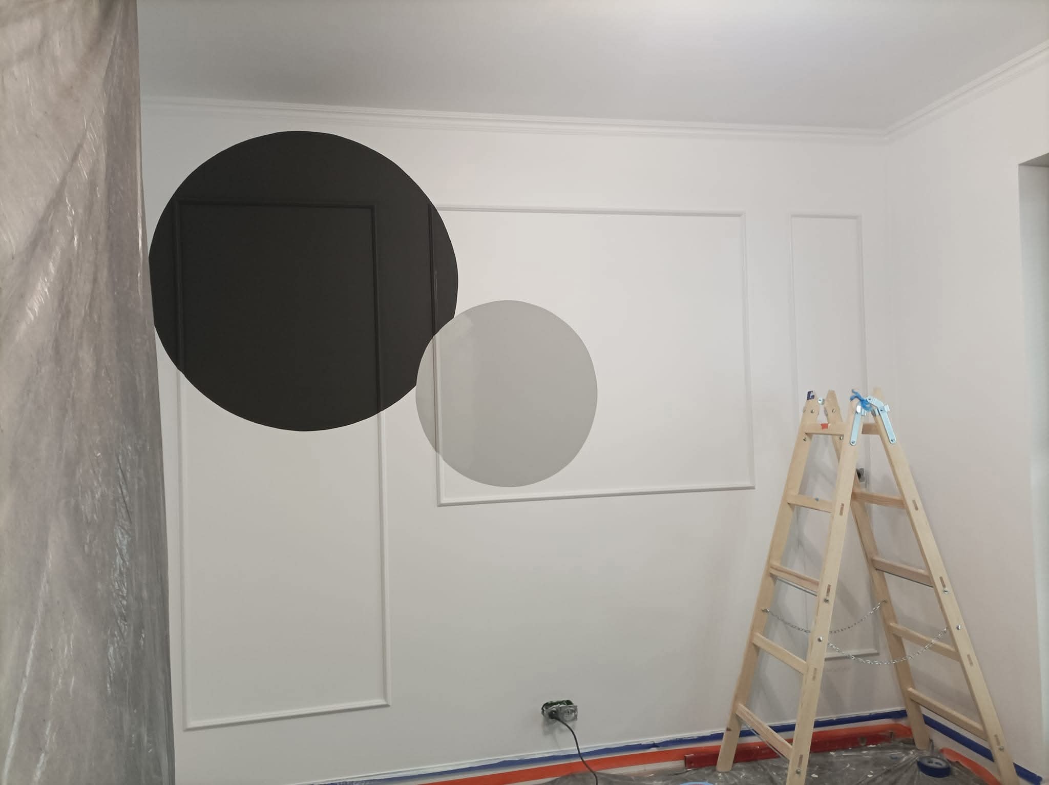 Ściana z dekoracyjnymi listwami i pomalowanymi okręgami w odcieniach szarości. Widoczna drabina i folia ochronna, sugerujące prace malarskie w toku.