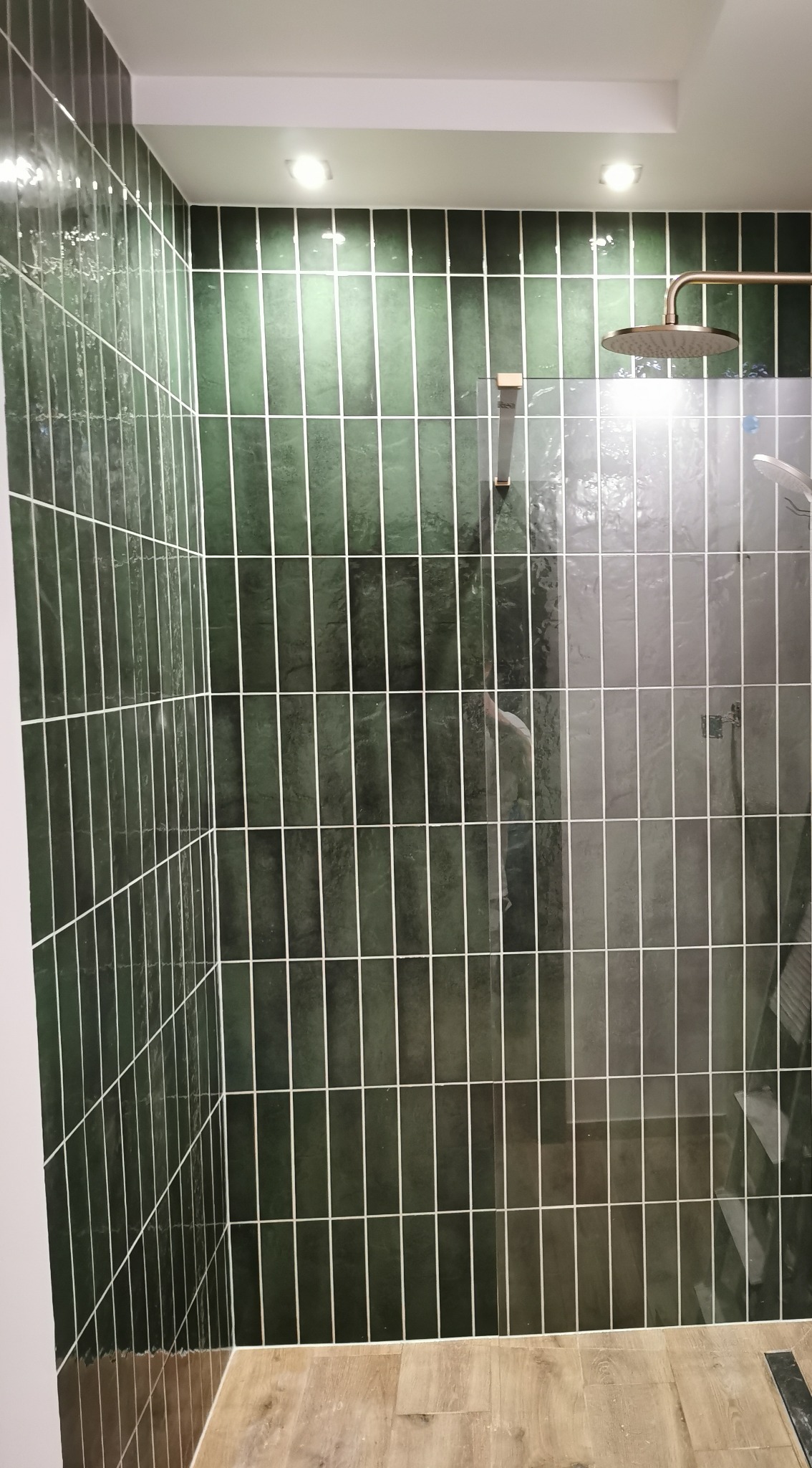 Wykończona kabina prysznicowa z zielonymi, podłużnymi płytkami i złotą armaturą. Widoczna szklana ściana kabiny i drewnopodobna podłoga. Nowoczesny design.