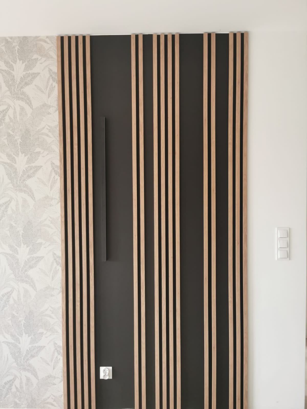 Panel ścienny z drewnianych listew na ciemnym tle, obok ściana z tapetą w liście i biała ściana z włącznikami. Nowoczesny design wnętrza.