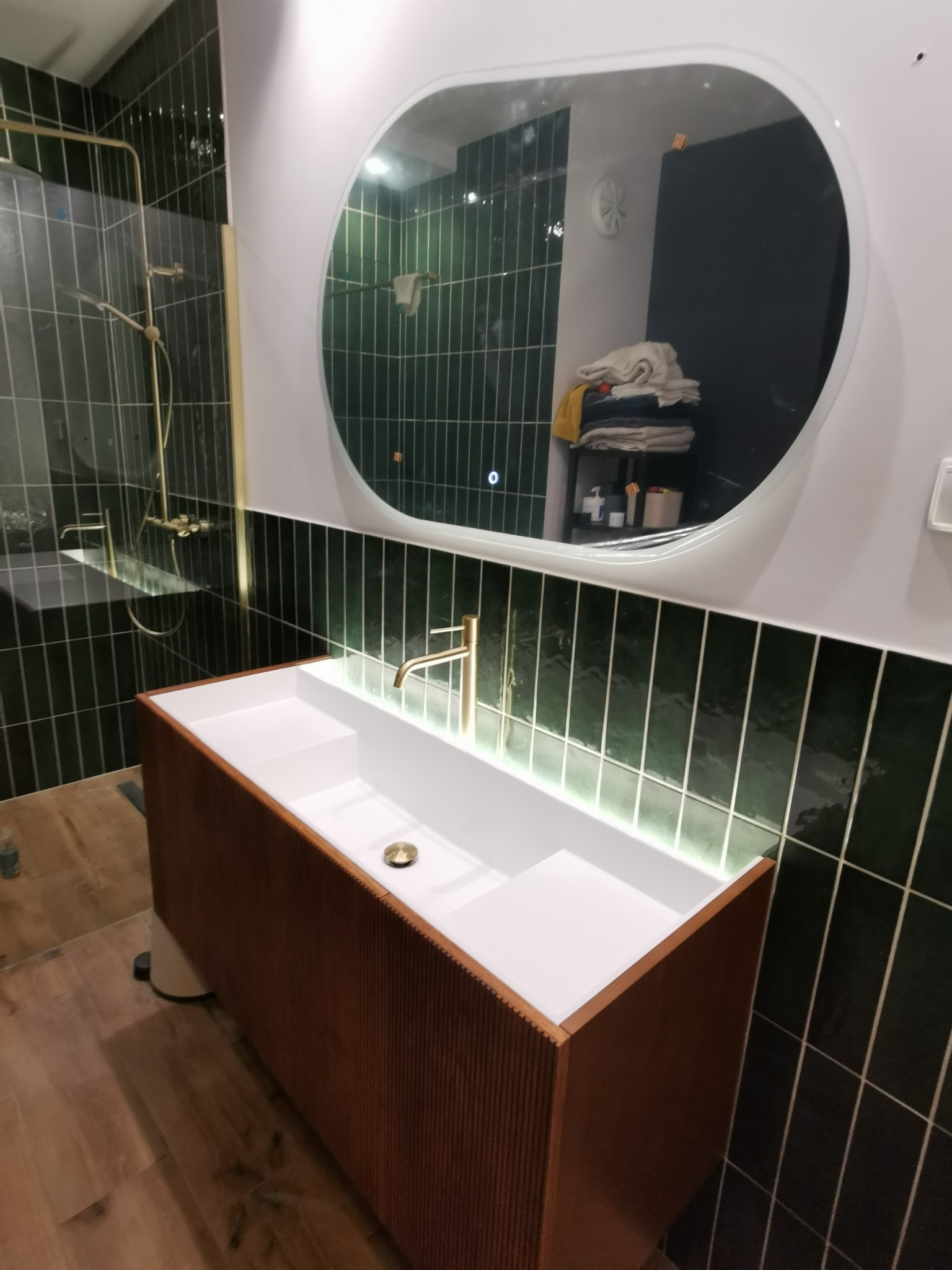 Nowoczesna łazienka z umywalką nablatową na drewnianej szafce, zielone płytki na ścianach i owalne lustro z podświetleniem LED. Złota armatura dodaje elegancji.