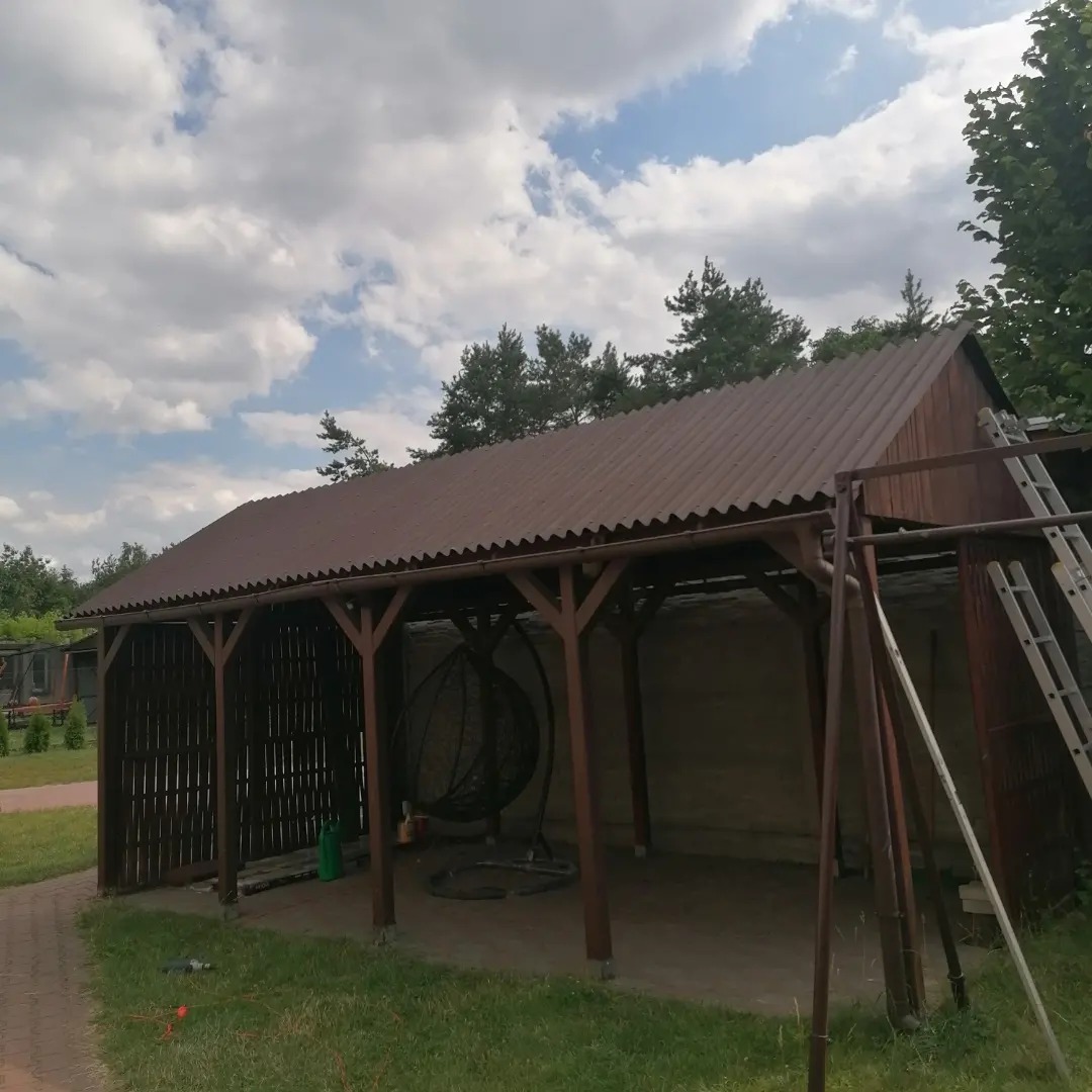 Zadaszona drewniana konstrukcja z brązowym falistym dachem, drabina oparta o bok, wiszące krzesło wewnątrz, widok z zewnątrz w słoneczny dzień.