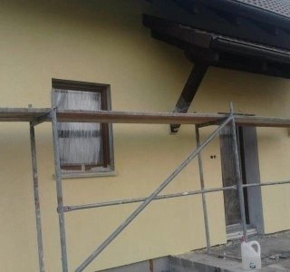 Elewacja budynku w trakcie ocieplania, widoczne rusztowanie, okno z zasłoniętą szybą, fragment dachu z rynną i plastikowy kanister.