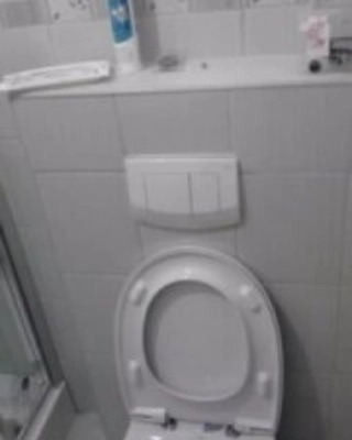 Biała toaleta z otwartą klapą i podwójnym przyciskiem spłukiwania na tle białych płytek w łazience, widoczna butelka kosmetyku na półce.