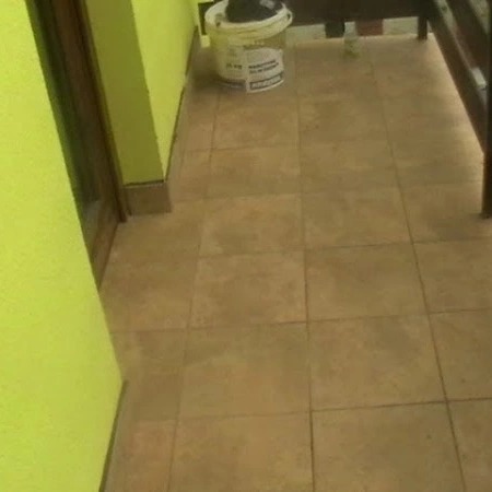 Balkon z brązowymi płytkami podłogowymi, widoczna ściana pomalowana na jasno zielono i wiadro z farbą w rogu.