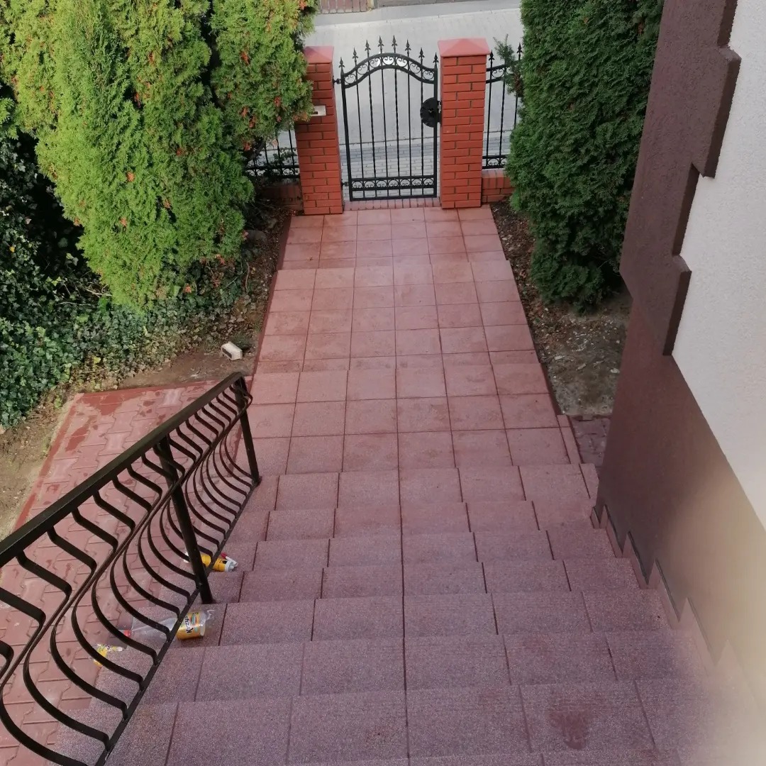 Czerwone płytki chodnikowe prowadzące po schodach do metalowej bramy z ceglanymi słupkami, otoczonej tujami.