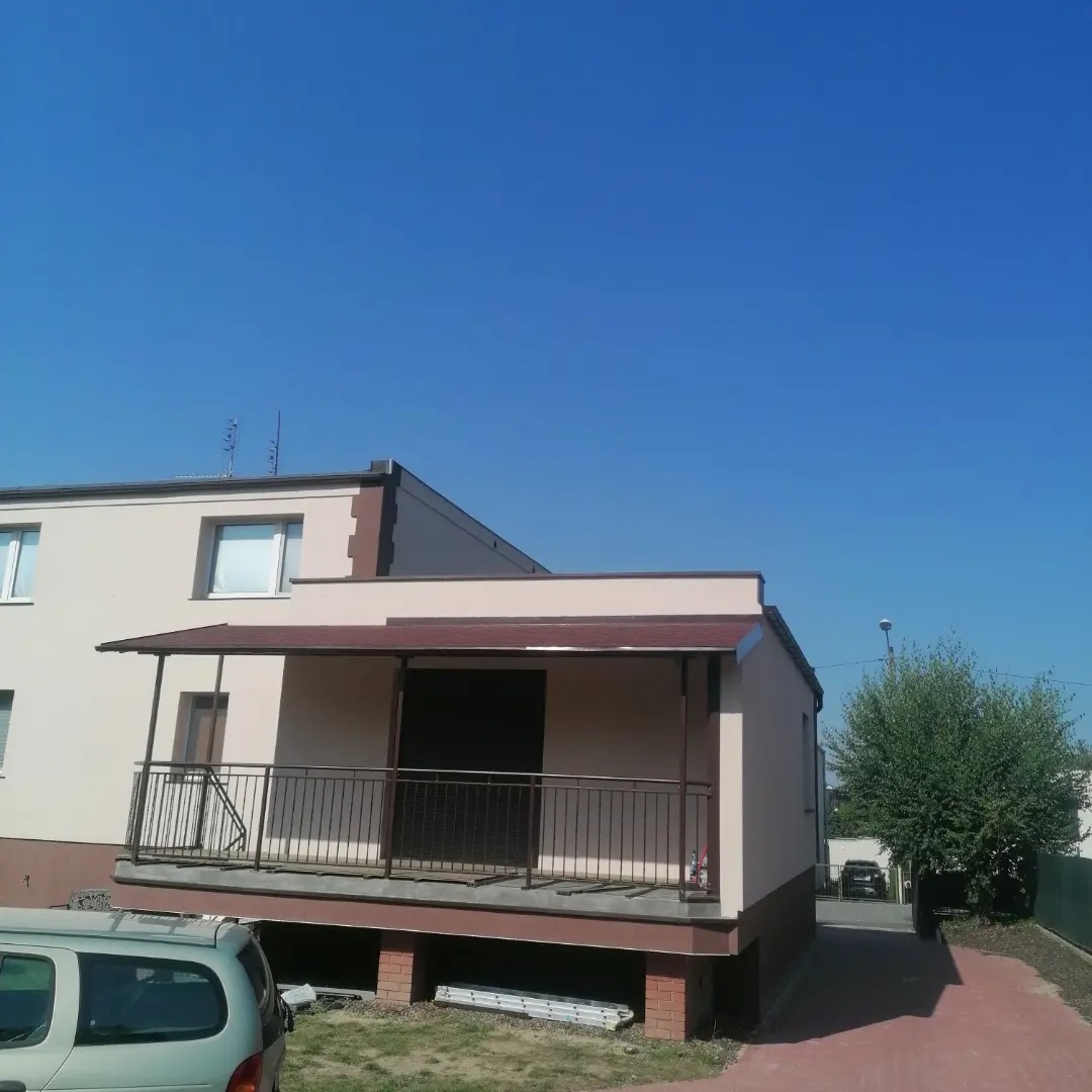 Dwukondygnacyjny budynek z beżową elewacją i brązowymi akcentami, z zadaszonym tarasem z metalową balustradą. Widoczny fragment samochodu na pierwszym planie i błękitne niebo w tle.