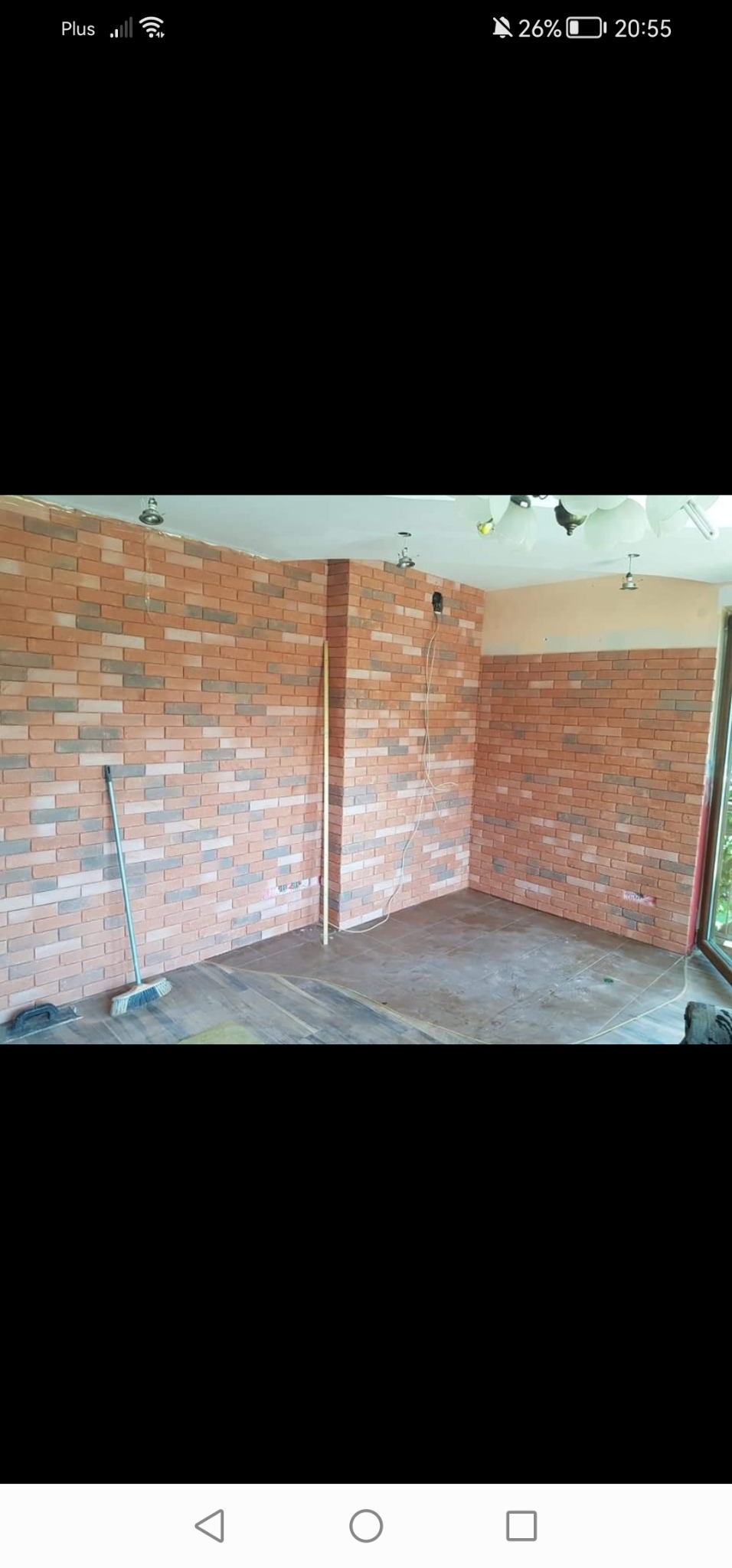 Wnętrze pokoju z częściowo wykończonymi ścianami z dekoracyjnej cegły w odcieniach czerwieni i szarości, widoczna drewnopodobna podłoga, listwa przypodłogowa oraz narzędzia: miotła i kielnia.