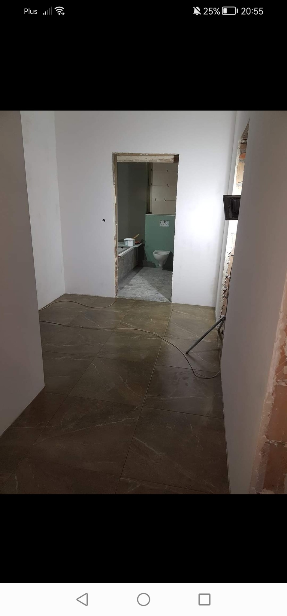 Wnętrze w trakcie remontu: brązowe płytki podłogowe z wzorem marmuru, surowe ściany przygotowane do malowania, widok na łazienkę z częściowo wykończoną instalacją sanitarną i oświetleniem roboczym.