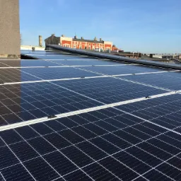 Rząd paneli słonecznych na dachu budynku w słoneczny dzień, z widokiem na sąsiednie budynki i błękitne niebo.