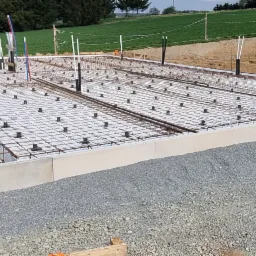 Zbrojona płyta fundamentowa z instalacjami pionowymi, przygotowana do wylewki betonu, widok na teren budowy.