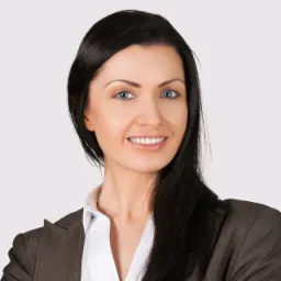 Portret uśmiechniętej kobiety w brązowej marynarce, reprezentującej wirtualne biuro w Krakowie, z charakterystycznym, lekkim uśmiechem i intensywnym spojrzeniem.