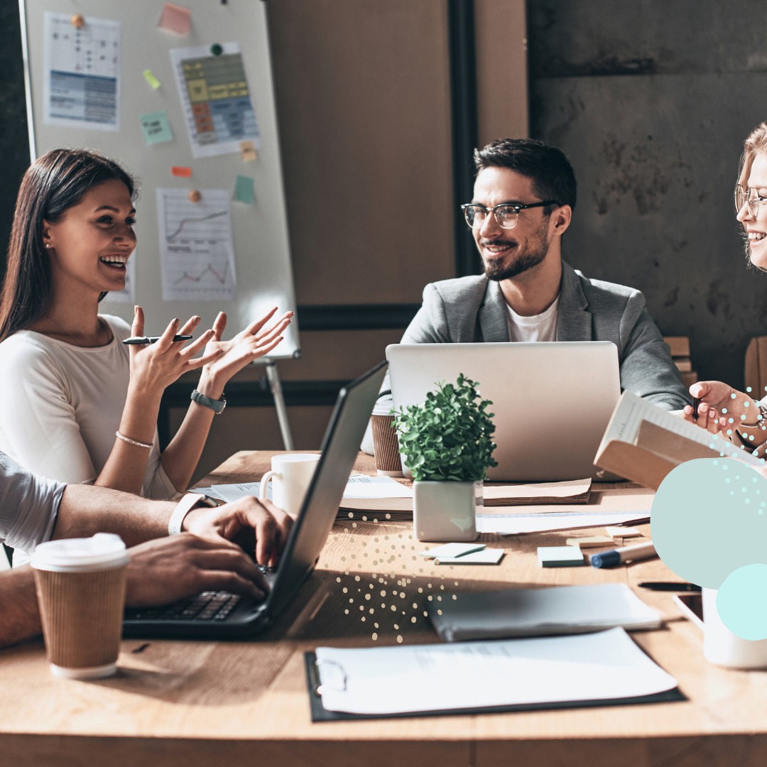 Zespół w biurze podczas burzy mózgów: kobieta gestykuluje z uśmiechem, mężczyzna pisze na laptopie, w tle flipchart z wykresami i dane, na stole kawa i roślina doniczkowa.