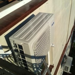 KlimaSort - Zewnętrzna jednostka klimatyzatora marki AUX zamontowana na ścianie budynku, widoczna z balkonu z metalową balustradą, z drabiną opartą o ścianę w pobliżu.