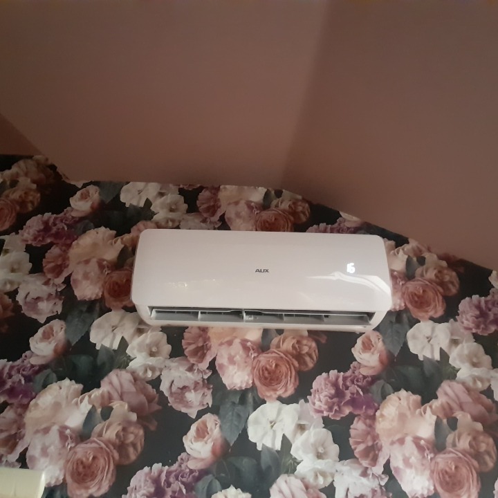 Biały klimatyzator marki AUX zamontowany na ścianie pokrytej tapetą w kwiaty, widoczny wyświetlacz z temperaturą.