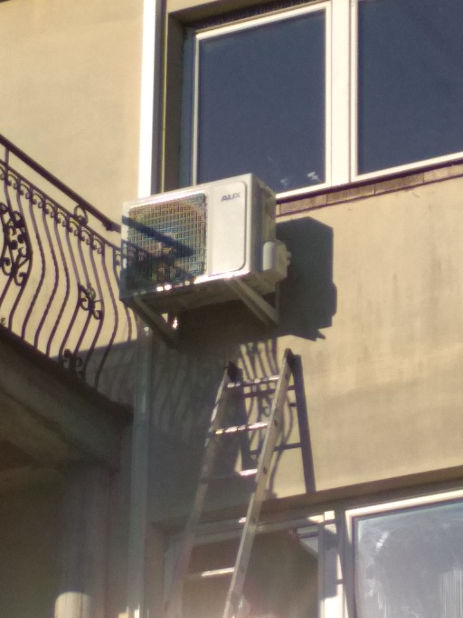 Zewnętrzna jednostka klimatyzacji AUX zamontowana na wsporniku na ścianie budynku, z drabiną opartą o ścianę poniżej.