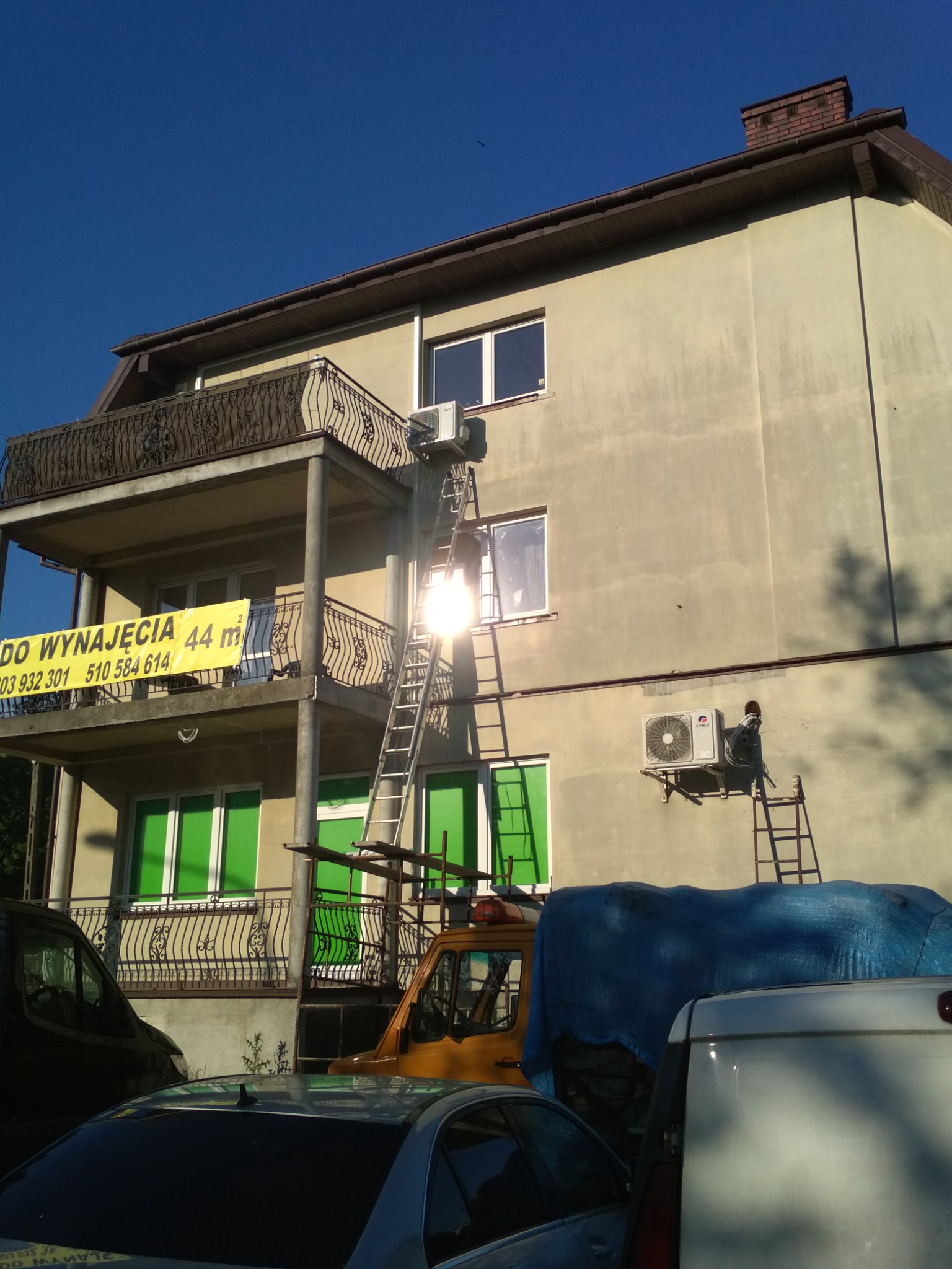 Montaż klimatyzatora na ścianie dwupiętrowego budynku z balkonami, pracownik na drabinie obok urządzenia, w tle błękitne niebo i zaparkowane samochody.