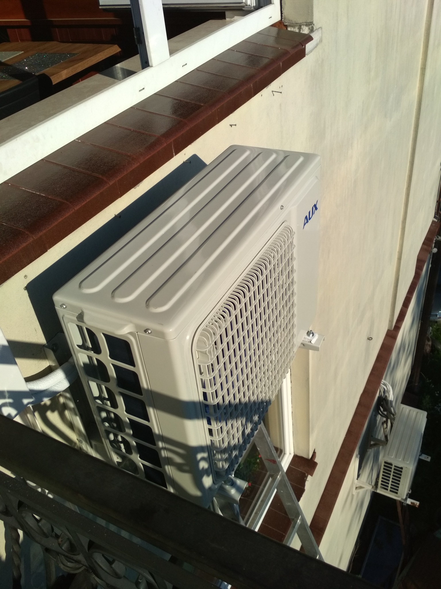 Zewnętrzna jednostka klimatyzatora marki AUX zamontowana na ścianie budynku, widoczna z balkonu z metalową balustradą, z drabiną opartą o ścianę w pobliżu.