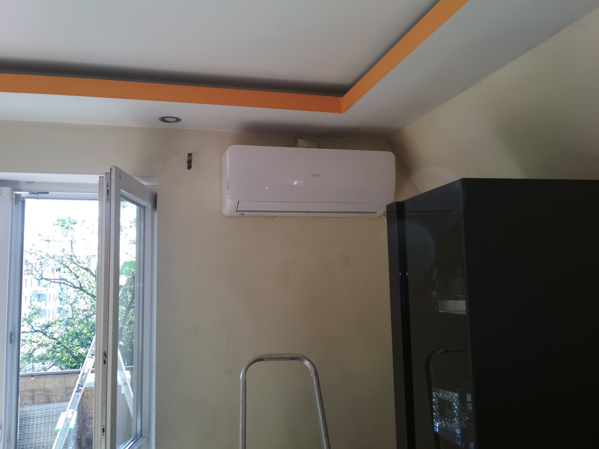 Biały klimatyzator marki AUX zamontowany na ścianie w jasnym pokoju, widoczna drabina aluminiowa i otwarte okno balkonowe z widokiem na zieleń.