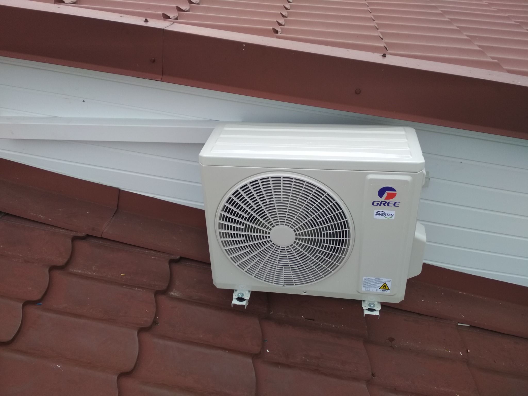 Zewnętrzna jednostka klimatyzatora marki Gree zamontowana na wspornikach na dachu pokrytym brązową dachówką, widoczna biała elewacja budynku.