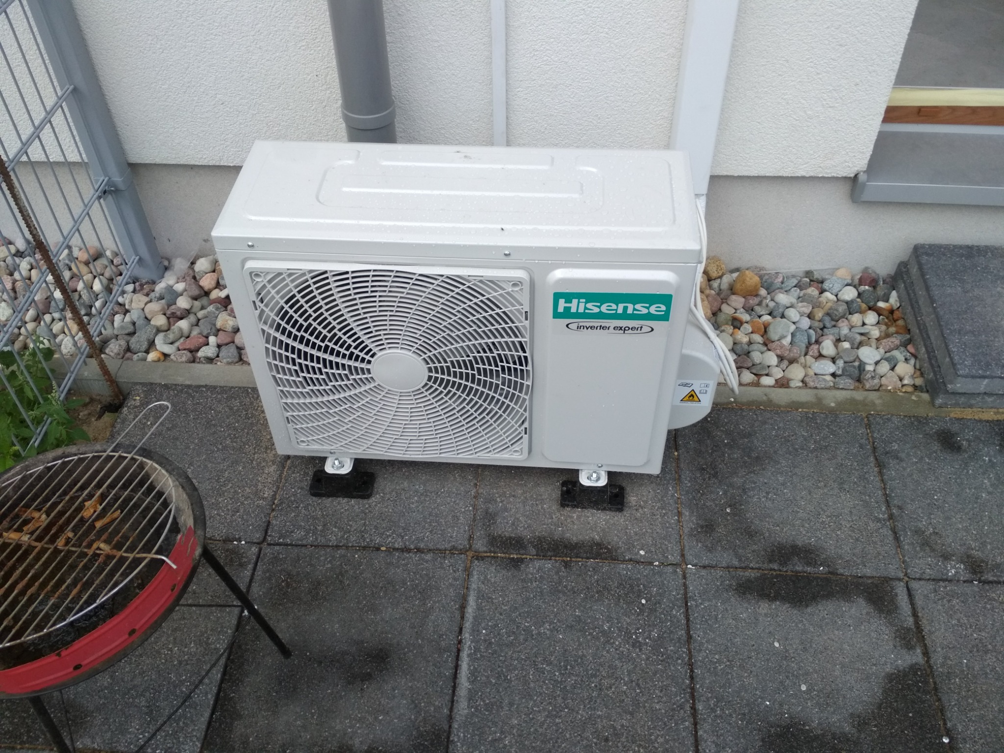 Zewnętrzna jednostka klimatyzatora Hisense Inverter Expert umieszczona na szarych płytach chodnikowych przy ścianie budynku, obok grilla z resztkami węgla.