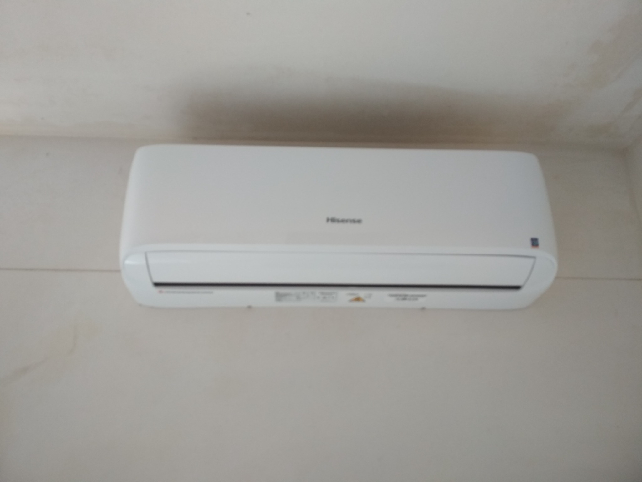 Biała jednostka klimatyzacyjna Hisense zamontowana na jasnej, lekko zabrudzonej ścianie w rogu pomieszczenia.