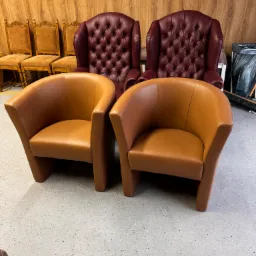 Dwa brązowe, skórzane fotele klubowe w Tarnowie, z dwoma bordowymi fotelami typu chesterfield w tle i rzędem drewnianych krzeseł za nimi.