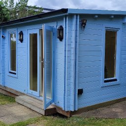 daz-build - Niebieski, drewniany domek letniskowy z otwartymi dwuskrzydłowymi drzwiami, oświetlony zewnętrznymi lampami, w otoczeniu zielonego trawnika i drzew.