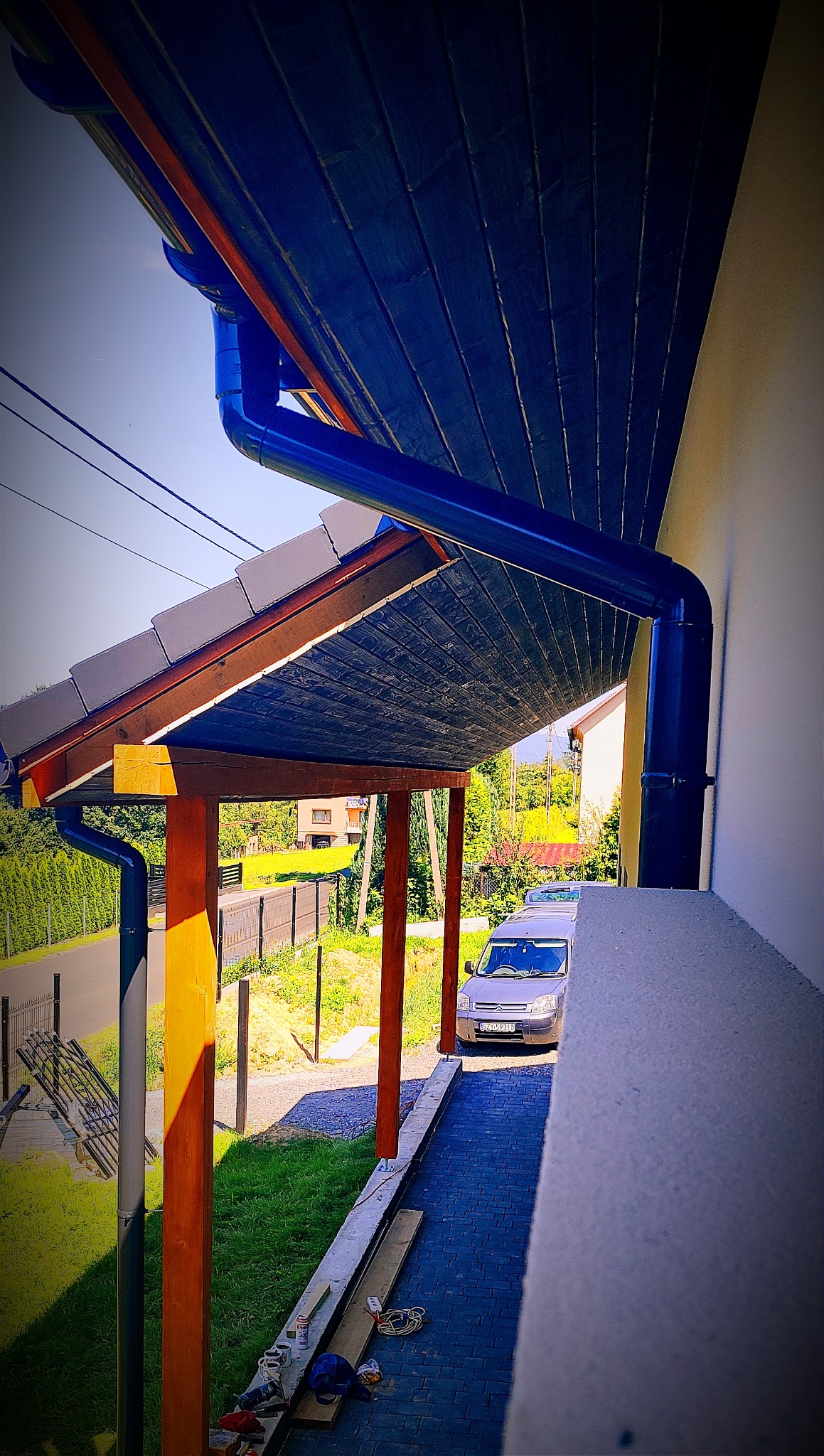 Zadaszenie tarasu z drewnianymi słupami, widoczne rynny, dachówka i podbitka, częściowo w trakcie budowy, narzędzia na podłożu.