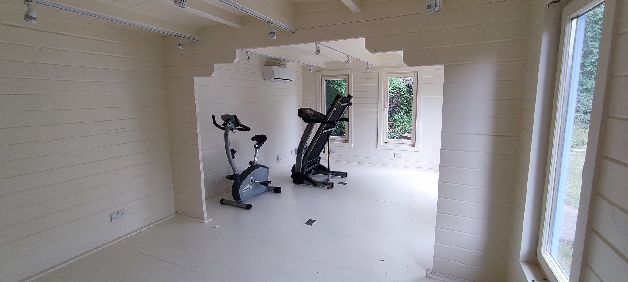 Wnętrze jasnego, drewnianego domku z oknami, zaaranżowanego na domową siłownię z rowerem stacjonarnym i bieżnią.