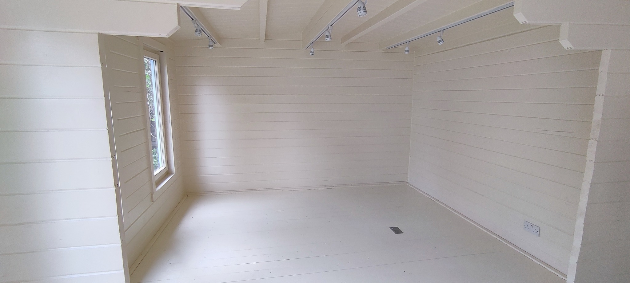 Wnętrze pomieszczenia wykończonego boazerią w jasnym odcieniu, z oknem i zamontowanym oświetleniem szynowym.