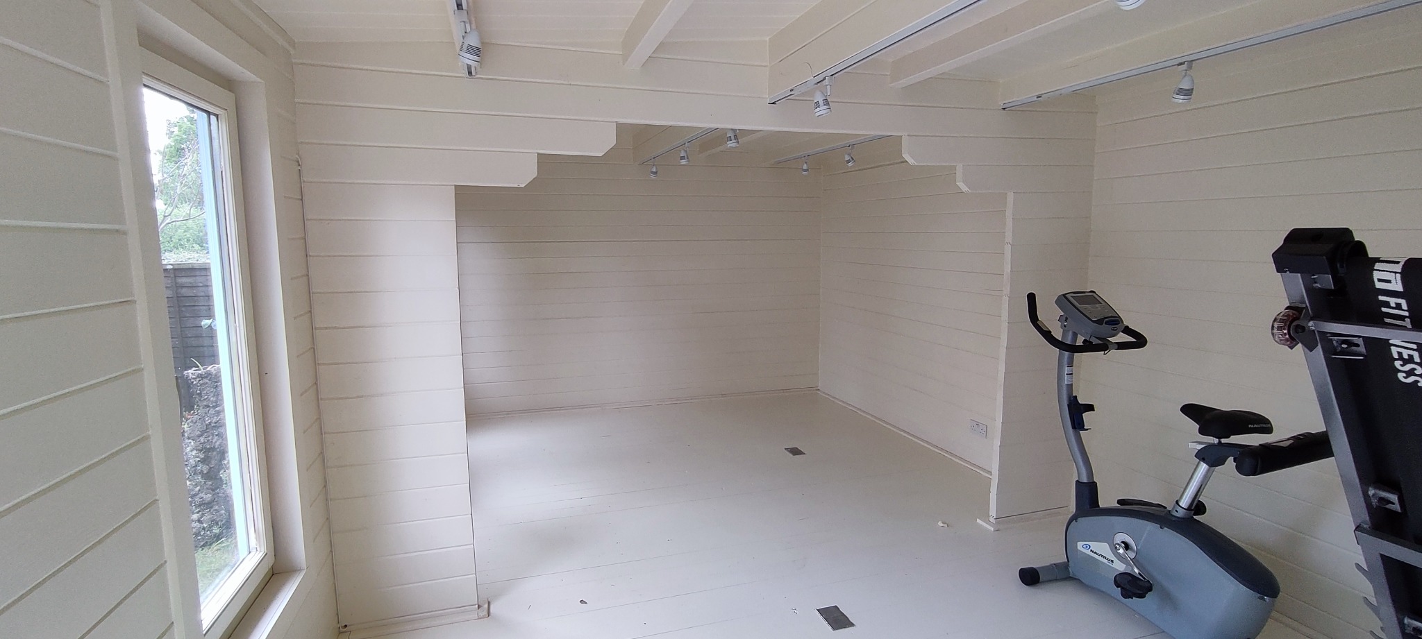 Świeżo ocieplone i pomalowane na kremowo wnętrze pomieszczenia z drewnianą boazerią, oświetleniem punktowym na szynoprzewodach i oknem, z widocznym sprzętem do ćwiczeń.