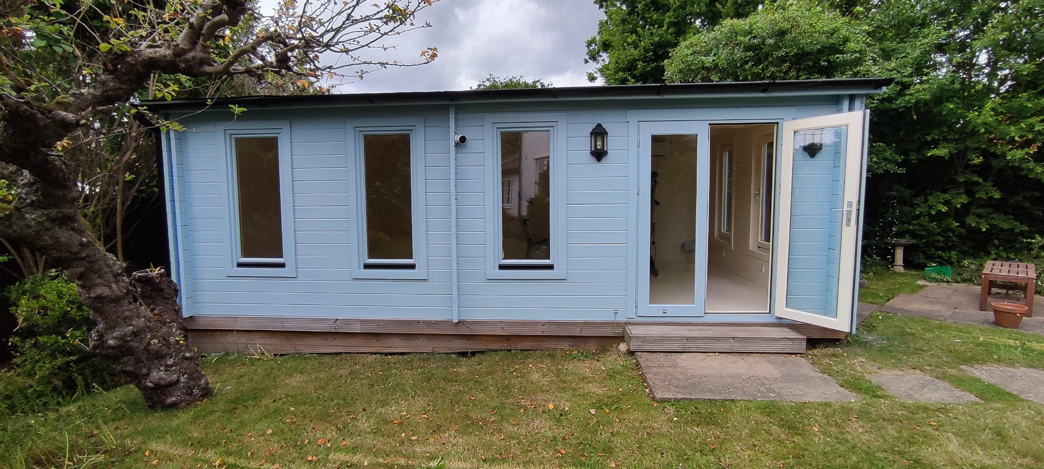 Niebieska drewniana altana ogrodowa z trzema oknami i otwartymi przeszklonymi drzwiami, widoczna lampa zewnętrzna, na tle zielonego ogrodu z drzewami.