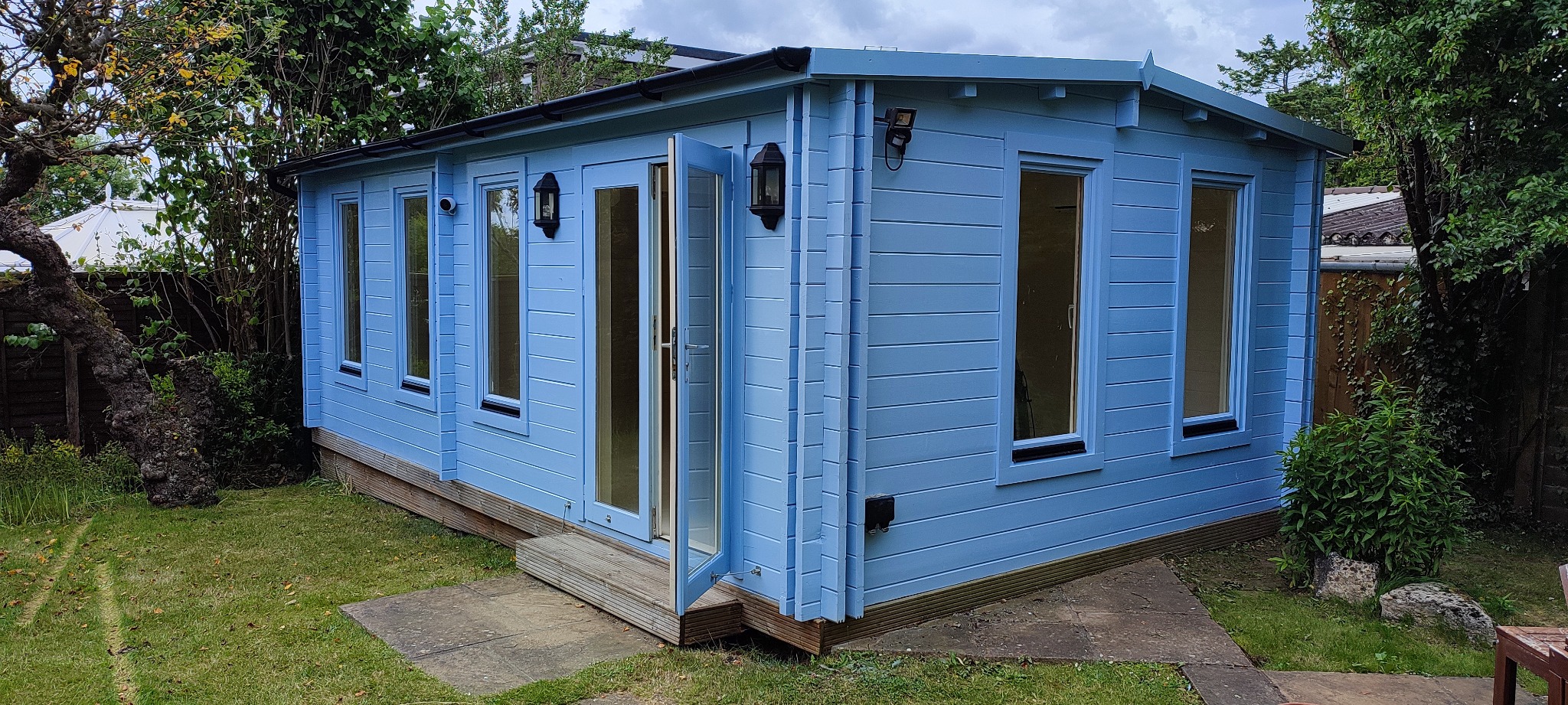 Niebieski, drewniany domek letniskowy z otwartymi dwuskrzydłowymi drzwiami, oświetlony zewnętrznymi lampami, w otoczeniu zielonego trawnika i drzew.