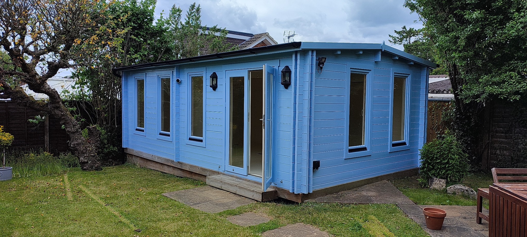 Niebieski domek ogrodowy z drewnianą elewacją, otwartymi drzwiami i oknami, oświetlony lampami zewnętrznymi, na tle zielonego trawnika i drzew