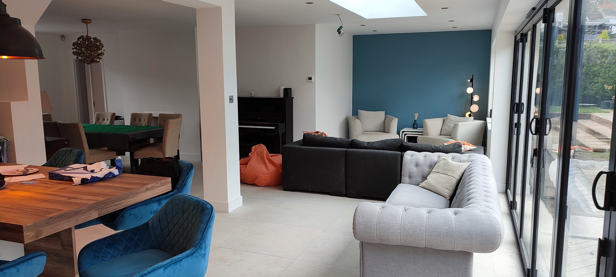 Jasne, przestronne wnętrze domu z nowoczesnymi meblami, w tym szarą sofą chesterfield, czarnym pianinem i drewnianym stołem z aksamitnymi krzesłami, oświetlone naturalnym światłem przez duże okna...