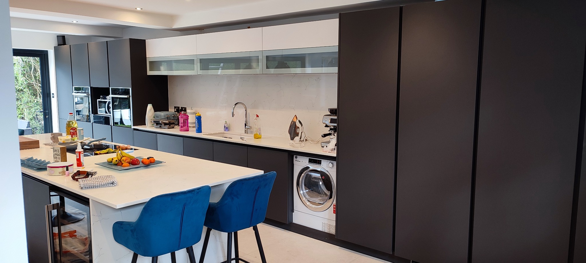 Nowoczesna kuchnia z ciemnymi szafkami, białym blatem i wyspą, z niebieskimi krzesłami barowymi, sprzęt AGD w zabudowie oraz pralka wkomponowana w zabudowę kuchenną.