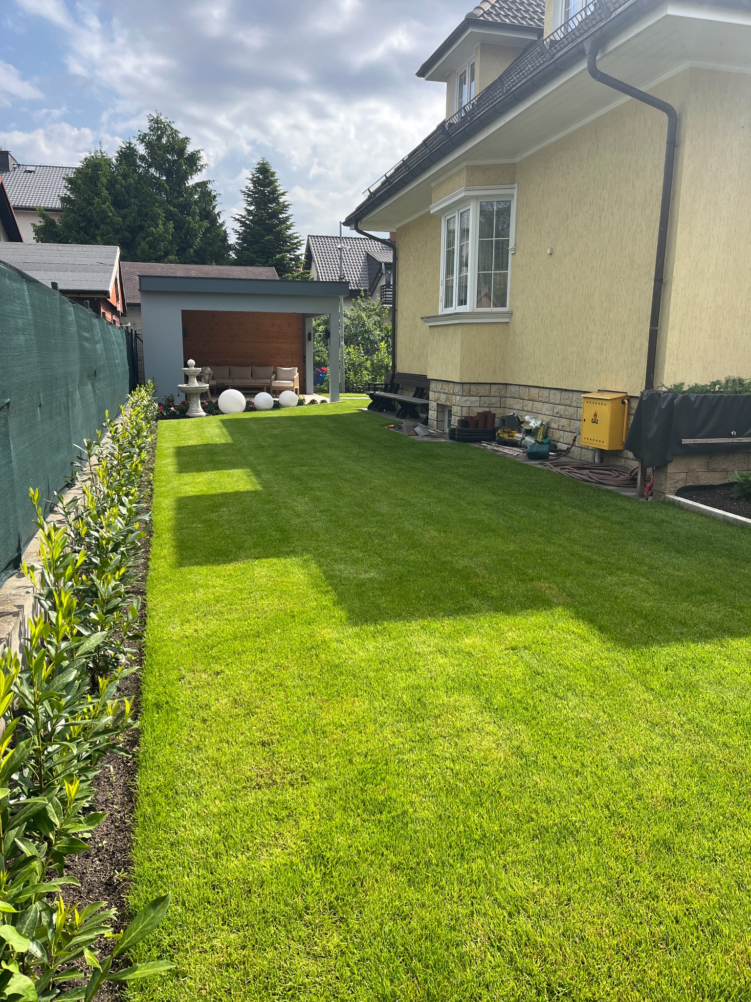 Perfekcyjnie skoszony trawnik przydomowy z równo przyciętym żywopłotem wzdłuż ogrodzenia, z widocznym domem i altaną w tle, w słoneczny dzień.