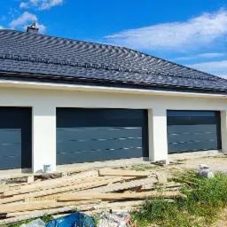 Nowoczesny garaż z trzema segmentowymi bramami w kolorze antracytowym, widok z zewnątrz, z żółtym samochodem dostawczym firmy zajmującej się stolarką okienną i drzwiową w tle, teren budowy...