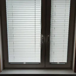 Dwu-skrzydłowe okno PCV z zamontowanymi, jasnymi plisami okiennymi, widoczny grzejnik pod oknem.