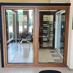 Ekspozycja stolarki okiennej z drewnopodobną ramą i czarną obudową, prezentowana w salonie sprzedaży z widocznym kalendarzem firmowym na ścianie.