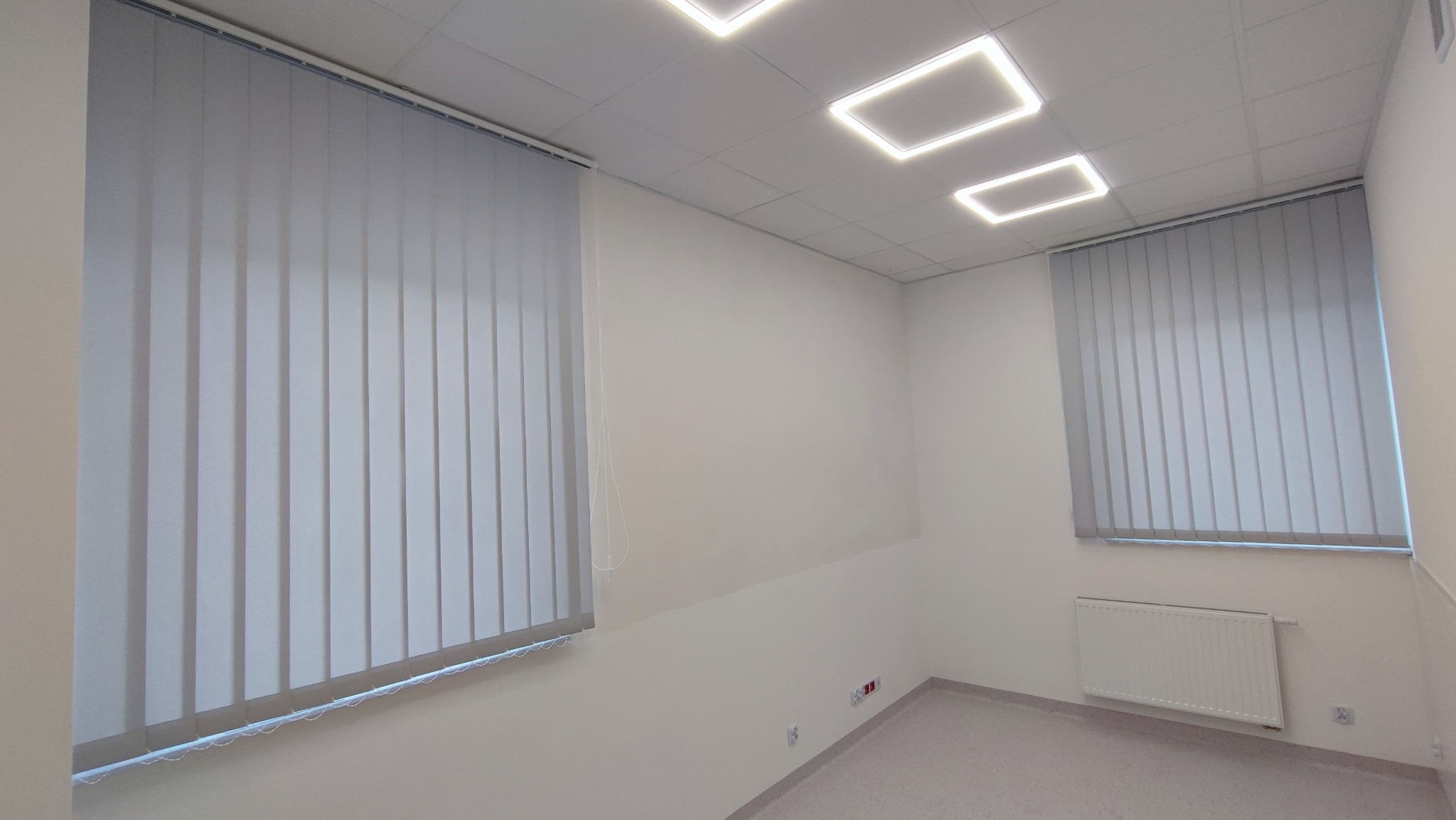 Wnętrze biurowe z dwoma oknami przesłoniętymi jasnoszarymi żaluzjami pionowymi (wertykalnymi), oświetlone kwadratowymi lampami sufitowymi LED.